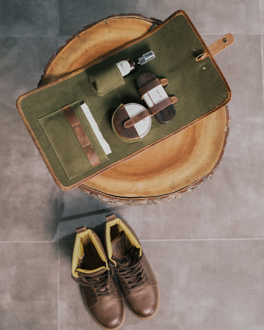 Green leather care kit bag with grooming products on a wooden block, brown boots on a tiled floor.