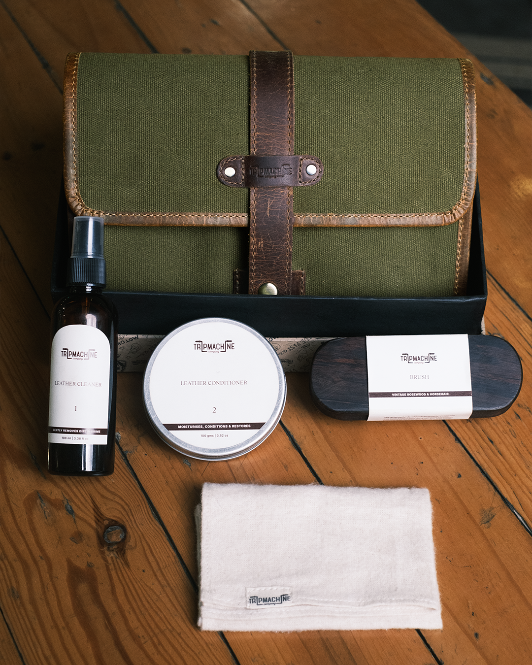Set of leather care kit products including a green leather case, cleaner, conditioner, and cloth on a wooden surface.